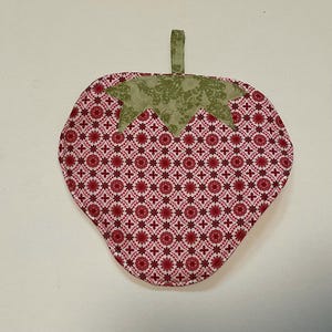 May include: A strawberry-shaped pot holder with a red and white floral pattern. The top of the strawberry is green with a leaf design, and it has a small green loop for hanging.