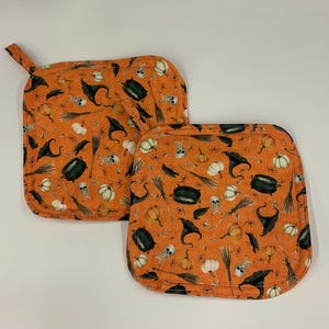 Puede incluir: Dos agarraderas de tela cuadradas con fondo naranja y un patrón repetitivo de sombreros de bruja negros, calderos, calaveras blancas, calabazas y escobas. Cada agarradera tiene esquinas redondeadas y una pequeña presilla de tela.