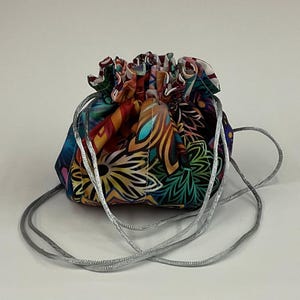 Puede incluir: Una pequeña bolsa con cordón con un vibrante estampado floral en tonos de azul, naranja, amarillo y verde. La bolsa tiene un cierre de cordón plateado y probablemente se utiliza para transportar objetos pequeños. La bolsa es de tela.