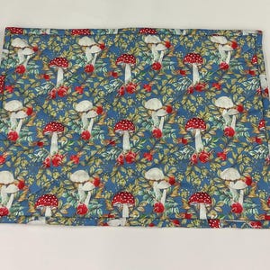 May include: A rectangular placemat with a repeating pattern of red-capped mushrooms, white stems, and red berries on a blue background. The placemat has a quilted texture and a stitched border. The design includes green leaves and yellow floral accents.