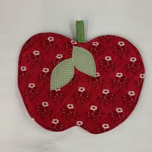 May include: Red apple-shaped pot holder with a green leaf design. The pot holder is made of fabric with a white floral pattern.