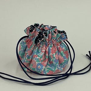Puede incluir: Una pequeña bolsa redonda con cordón y un estampado floral en rosa, azul y verde. La bolsa tiene un forro de lunares blancos y negros y un cordón azul marino. La bolsa probablemente se utiliza para guardar objetos pequeños.