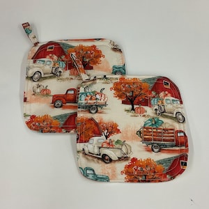 May include: Two square fabric pot holders featuring a repeating autumn-themed print. The design includes red barns, vintage pickup trucks in white and red, orange fall foliage, and various pumpkins. Each pot holder has a small fabric hanging loop.