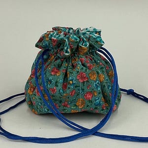 May include: A small, round drawstring pouch with a teal background and a floral pattern in shades of pink, orange, and blue. The pouch has a gathered top and is secured with a bright blue drawstring.