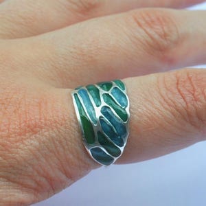 May include: A silver ring with a green and blue enamel design. The ring is on a finger, showcasing the intricate pattern of the enamel. The ring's design features a series of interconnected shapes in shades of green and blue.