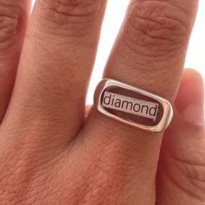 May include: A silver ring with the word "diamond" in a rectangular setting. The ring is worn on a finger, showcasing the text against a brown background within the setting. The ring is a simple, modern design.