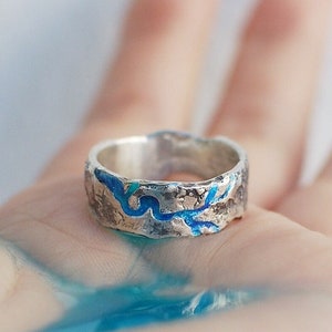 May include: A silver ring with a textured surface and a blue enamel design. The ring is on a hand.