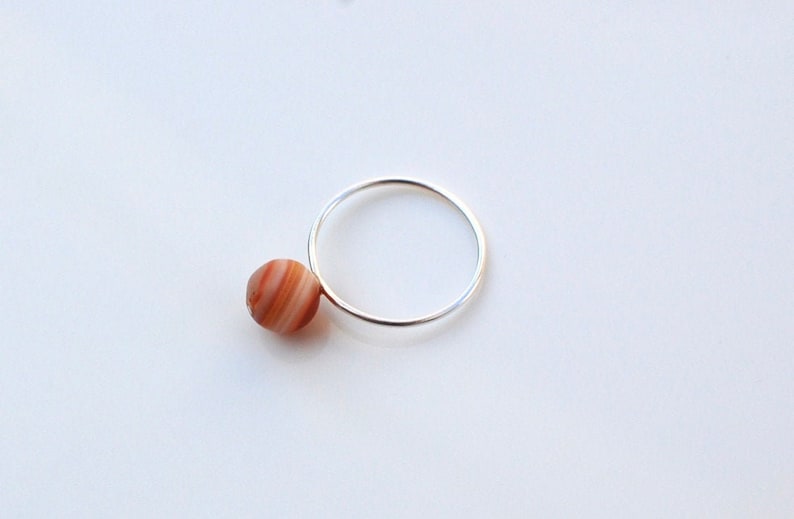 Puede incluir: Un anillo de plata con una sola piedra de &aacute;gata redonda, naranja y blanca unida a la banda.