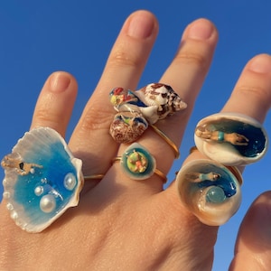 May include: A hand displays five gold-toned rings with seashell designs. Each ring features a unique miniature scene, including swimmers in blue water, pearls, and colorful shell details. The background is a clear blue sky.