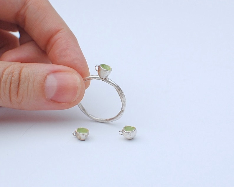 Peut inclure: Une bague en argent avec un petit charme en forme de tasse verte. Deux autres charmes en forme de tasse verte sont pr&eacute;sent&eacute;s sur une surface blanche.