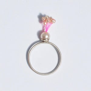 May include: A silver ring featuring a large pearl and a cluster of small, pale beads, connected by pink thread. The ring is circular and rests against a white backdrop. The beads are grouped, forming a delicate, feminine design, suitable for jewellery.