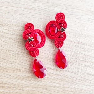 Könnte beinhalten: Ein Paar rote Ohrhänger mit einem Stern-Anhänger und einem roten Kristall-Tropfen. Die Ohrringe sind aus rotem Faden gefertigt und haben ein einzigartiges, filigranes Design.