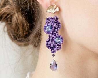 May include: Dangle earrings featuring a gold-toned ginkgo leaf stud, purple soutache cord embroidery arranged in swirling patterns, two round light purple crystals, and a single teardrop-shaped purple crystal pendant hanging at the base.