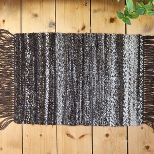 May include: A rectangular, hand-woven rug with a striped pattern in shades of brown, grey, and white. The rug has a fringe on both short ends. The rug is placed on a wooden surface.