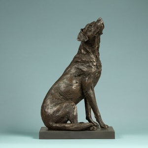 Puede incluir: Escultura de bronce de un perro sentado, mirando hacia arriba. El perro tiene una superficie texturizada y está montado sobre una base rectangular. La escultura está sobre un fondo azul claro. Objeto decorativo.