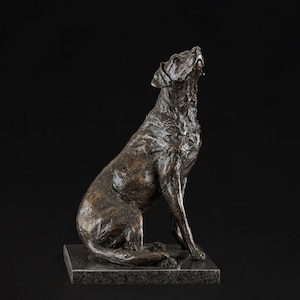 Puede incluir: Escultura de bronce de un perro Labrador Retriever sentado sobre una base negra. El perro está mirando hacia arriba y tiene una apariencia detallada y realista.