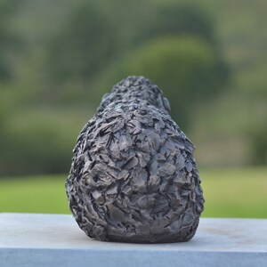 Puede incluir: Una escultura de bronce oscuro con una superficie texturizada, posiblemente representando una forma estilizada. La escultura est&aacute; colocada sobre una base rectangular gris claro, con un fondo verde borroso.