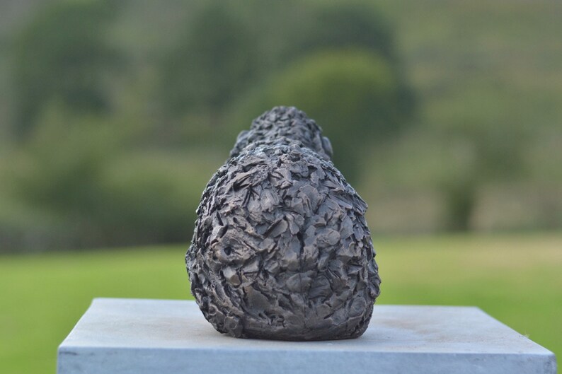 Puede incluir: Una escultura de bronce oscuro con una superficie texturizada, posiblemente representando una forma estilizada. La escultura est&aacute; colocada sobre una base rectangular gris claro, con un fondo verde borroso.