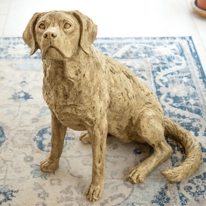 Puede incluir: Una detallada escultura de perro de color canela en posición sentada. El perro está colocado sobre una alfombra estampada con diseños florales azules y blancos. La escultura tiene una superficie texturizada, capturando la semejanza de un Labrador Retriever.