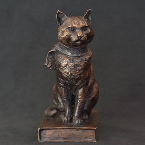 May include: A bronze cat statue sitting on a book. The cat wears a collar with a medallion and a cape. The sculpture has a detailed texture and a dark brown patina. The cat is posed in a regal, upright position.