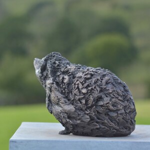 Puede incluir: Una escultura texturizada de color gris oscuro de un animal, posiblemente un oso o un erizo, se encuentra sobre una superficie gris claro. La escultura tiene una superficie rugosa y detallada y se encuentra sobre un fondo verde borroso, lo que sugiere un entorno al aire libre.