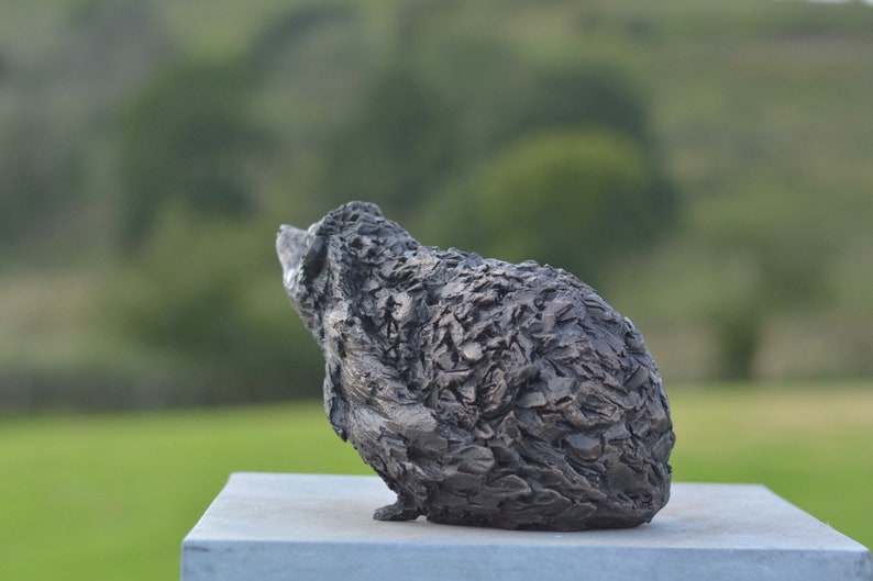 Puede incluir: Una escultura texturizada de color gris oscuro de un animal, posiblemente un oso o un erizo, se encuentra sobre una superficie gris claro. La escultura tiene una superficie rugosa y detallada y se encuentra sobre un fondo verde borroso, lo que sugiere un entorno al aire libre.