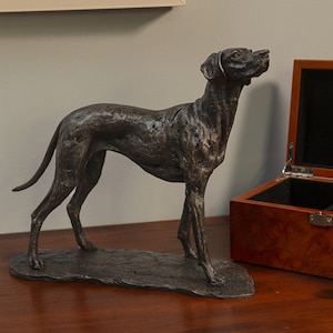Op de afbeelding: Een bronzen beeld van een hond die op een voetstuk staat. De hond kijkt naar rechts en heeft zijn hoofd omhoog gekanteld. Het beeld staat op een houten oppervlak.