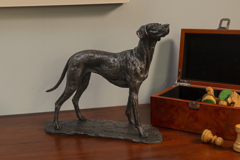 Op de afbeelding: Een bronzen beeld van een hond die op een voetstuk staat. De hond kijkt naar rechts en heeft zijn hoofd omhoog gekanteld. Het beeld staat op een houten oppervlak.