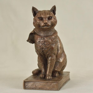 May include: A bronze cat statue sitting on a book. The cat has a decorative collar and a detailed chest design. The sculpture is a warm brown colour and has a detailed texture.