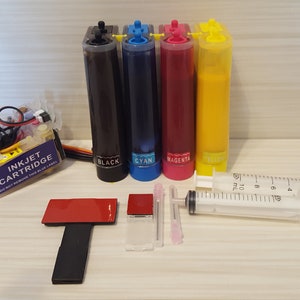 May include: A set of four ink cartridges for a printer, in black, cyan, magenta, and yellow. The cartridges are in clear plastic containers with labels. There are also two syringes, a red adhesive pad, and a clear plastic piece.