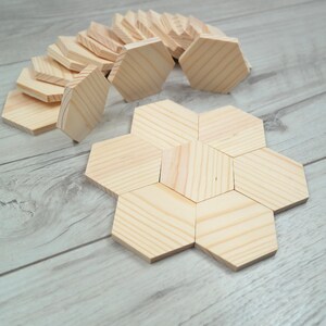 May include: A collection of light-colored, unfinished wooden hexagon tiles. One tile is placed in the center, surrounded by six identical tiles to form a larger hexagonal shape. Several additional loose wooden hexagon tiles are stacked behind.