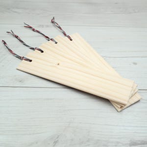 May include: A stack of unfinished wooden bookmarks. Each bookmark is rectangular and made of light-coloured wood. The bookmarks have holes at the top for string. The strings are black, red, and white.