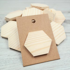 May include: A collection of unfinished light-coloured wooden hexagon shapes. One hexagon is attached to a rectangular brown cardboard display card with a circular hole at the top, while several other identical wooden hexagons are stacked behind it.