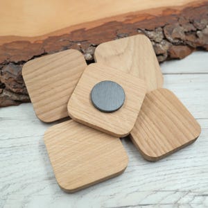 Könnte beinhalten: Ein Set aus vier quadratischen, hellbraunen Holzteilen mit abgerundeten Ecken. Jedes Teil weist eine glatte, natürliche Holzmaserung auf. Eines der Teile hat eine kreisförmige, dunkelgraue Magnetscheibe in der Mitte der Oberfläche.