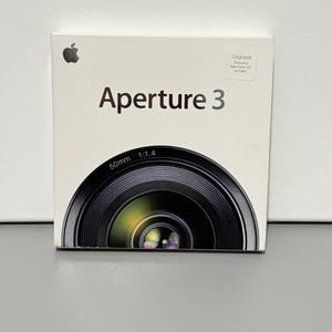 May include: Aperture 3 software box with an Apple logo. The box is white with the product name in a large, bold font. A detailed illustration of a camera lens is featured at the bottom, with the specifications 50mm 1:1.4.