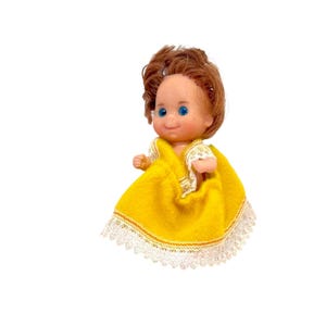 May include: A small doll with brown hair and blue eyes, wearing a yellow dress with white lace trim. The doll has a smiling expression and is set against a white background.