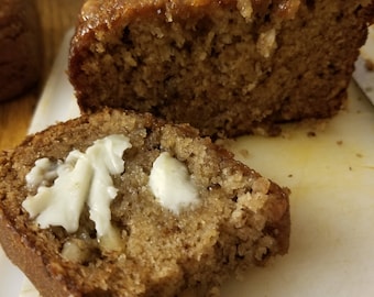 May include: A close-up of a loaf of banana bread with a slice cut out and buttered. The bread is light brown and has a slightly crumbly texture.