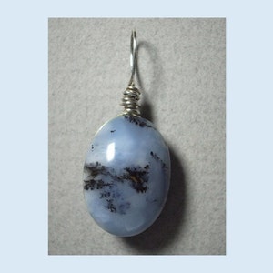 May include: A light blue oval pendant with a silver wire wrap and bail. The stone has dark brown and black inclusions, creating a unique pattern. The pendant is a jewelry item.