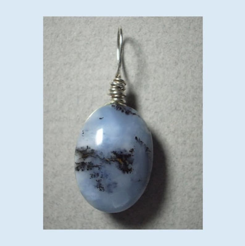 May include: A light blue oval pendant with a silver wire wrap and bail. The stone has dark brown and black inclusions, creating a unique pattern. The pendant is a jewelry item.