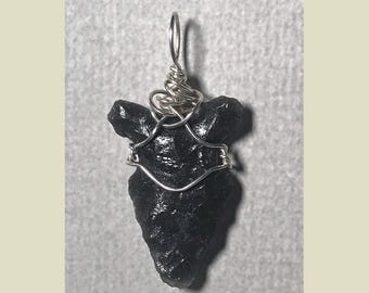 Puede incluir: Un colgante negro de forma irregular con un alambre de plata. El alambre está enrollado en la parte superior para formar un bucle y envuelve la piedra. El colgante está sobre un fondo gris.