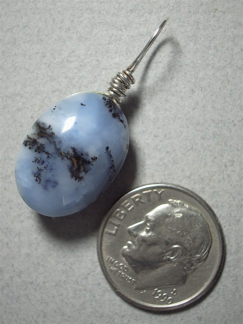 May include: A light blue dendritic opal pendant with a silver wire wrap. The oval-shaped stone features black and brown inclusions. A U.S. dime is next to the pendant for size reference.