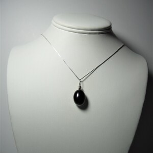May include: A silver necklace with a dark oval pendant displayed on a white mannequin. The pendant is a smooth, polished black stone, suspended from a delicate silver chain. The necklace is presented against a plain white background.