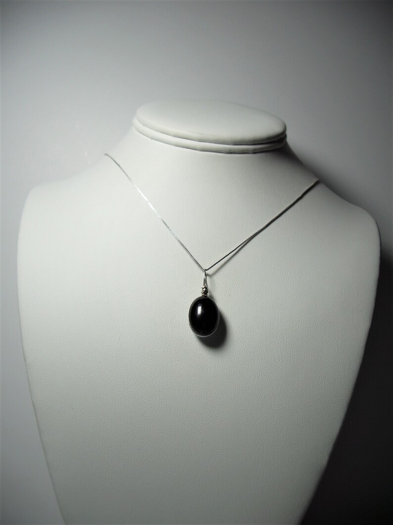 May include: A silver necklace with a dark oval pendant displayed on a white mannequin. The pendant is a smooth, polished black stone, suspended from a delicate silver chain. The necklace is presented against a plain white background.