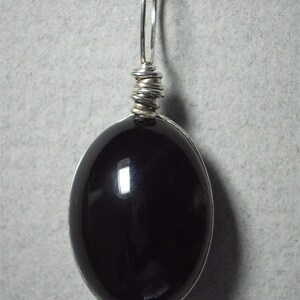 May include: A polished, oval-shaped black gemstone pendant with a silver wire wrap and bail. The pendant is suspended from a silver loop, creating a simple yet elegant design. The gemstone reflects light, showcasing its smooth surface.