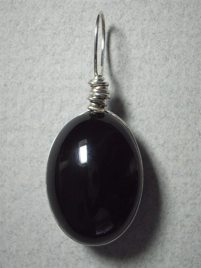 May include: A polished, oval-shaped black gemstone pendant with a silver wire wrap and bail. The pendant is suspended from a silver loop, creating a simple yet elegant design. The gemstone reflects light, showcasing its smooth surface.
