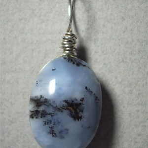 May include: Oval-shaped pendant with a light blue stone, featuring dark, tree-like inclusions. The stone is wire-wrapped with silver-colored metal and has a loop for a chain. The pendant is set against a neutral background.