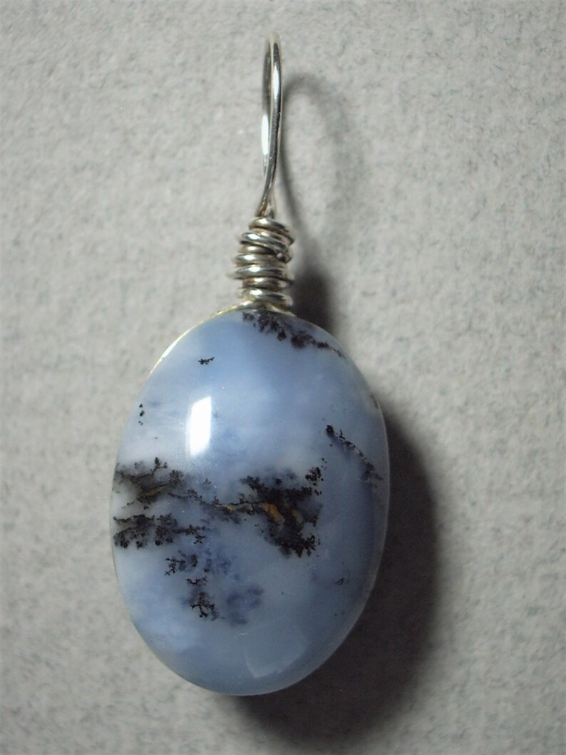 May include: Oval-shaped pendant with a light blue stone, featuring dark, tree-like inclusions. The stone is wire-wrapped with silver-colored metal and has a loop for a chain. The pendant is set against a neutral background.