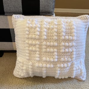 May include: White crocheted pillow with a textured, puffy design. The pillow is sitting on a black and white checkered surface.