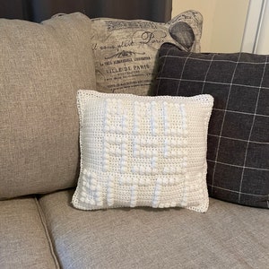 May include: White crocheted pillow with the word "CHEER" spelled out in raised stitches. The pillow is sitting on a brown couch with other pillows behind it.