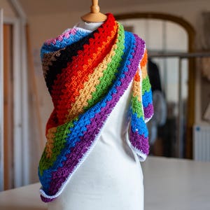 May include: A triangular crochet shawl featuring a vibrant rainbow colour palette with stripes of red, orange, yellow, green, blue, and purple. The shawl is crafted with a textured stitch pattern and finished with a clean white border along the edges.